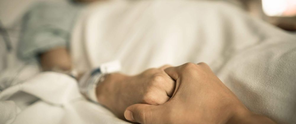 a person laying in a bed, another person out of the frame is holding his hand