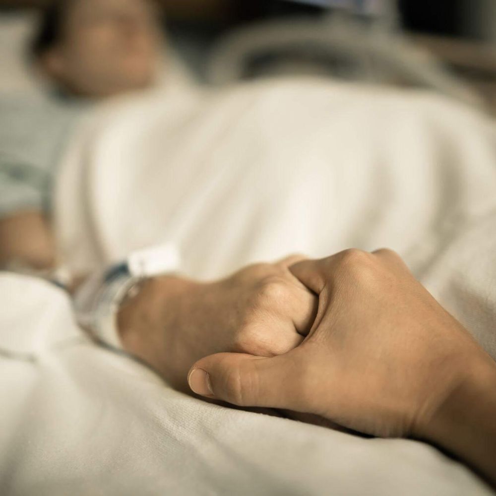 a person laying in a bed, another person out of the frame is holding his hand