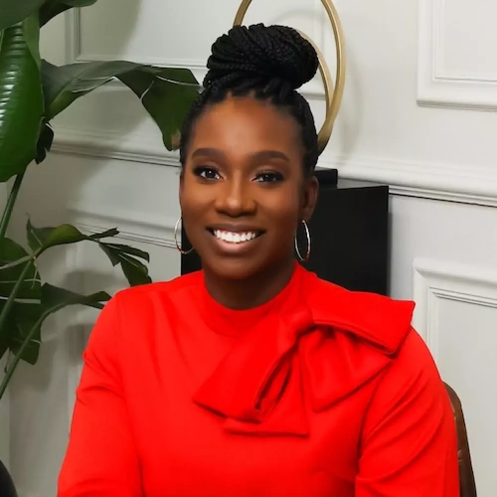 Smiling woman in a red blouse with a bow, sitting indoors with plants in the background.