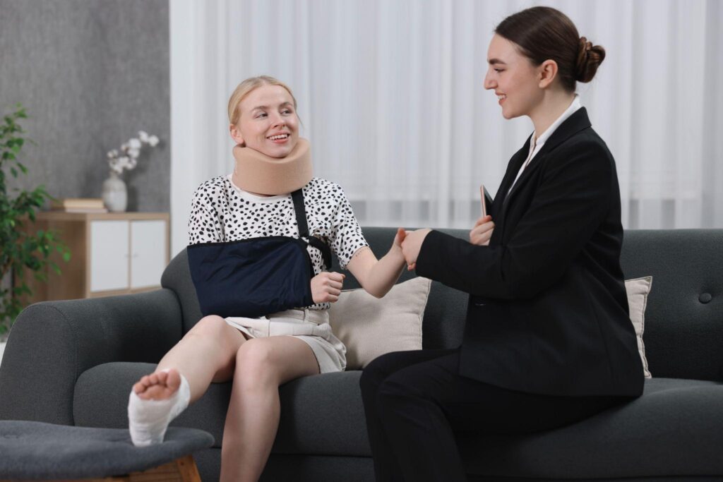 – The TD Injury Firm A woman with a neck brace, arm sling, and bandaged foot sits on a sofa, smiling while shaking hands with another woman in a suit in a living room.