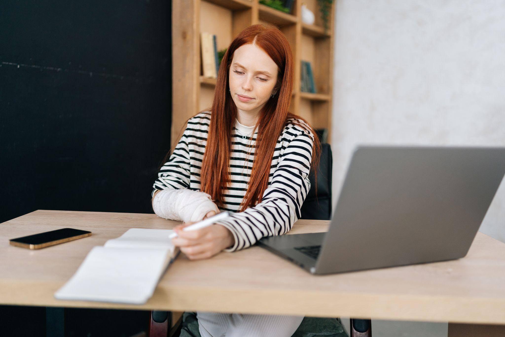 a woman sitting at a table with a laptop and taking notes on a notebook