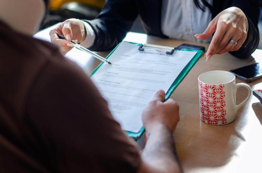 Two people discussing a document over coffee, with a pen pointing at a clipboard paper on a table.